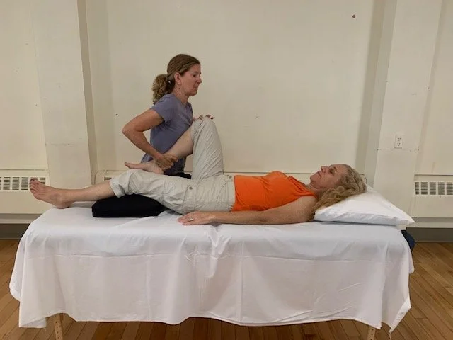A physical therapist assisting a woman lying on a massage table, performing a leg stretch exercise indoors.
